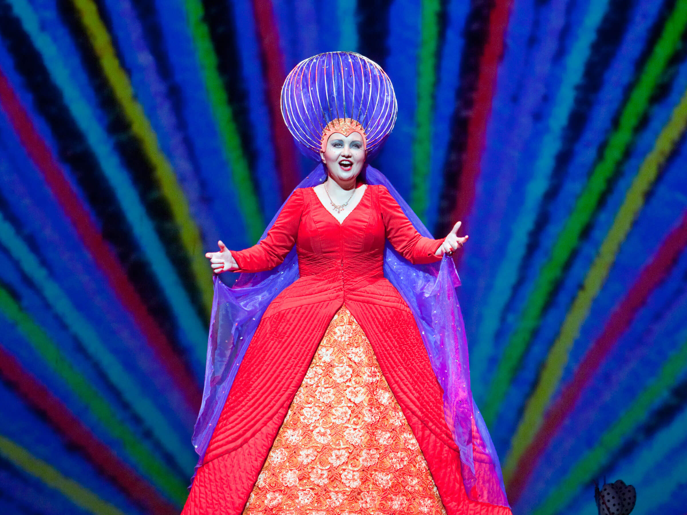 The Magic Flute character in a red dress and large purple headdress singing in front of a multicolored background