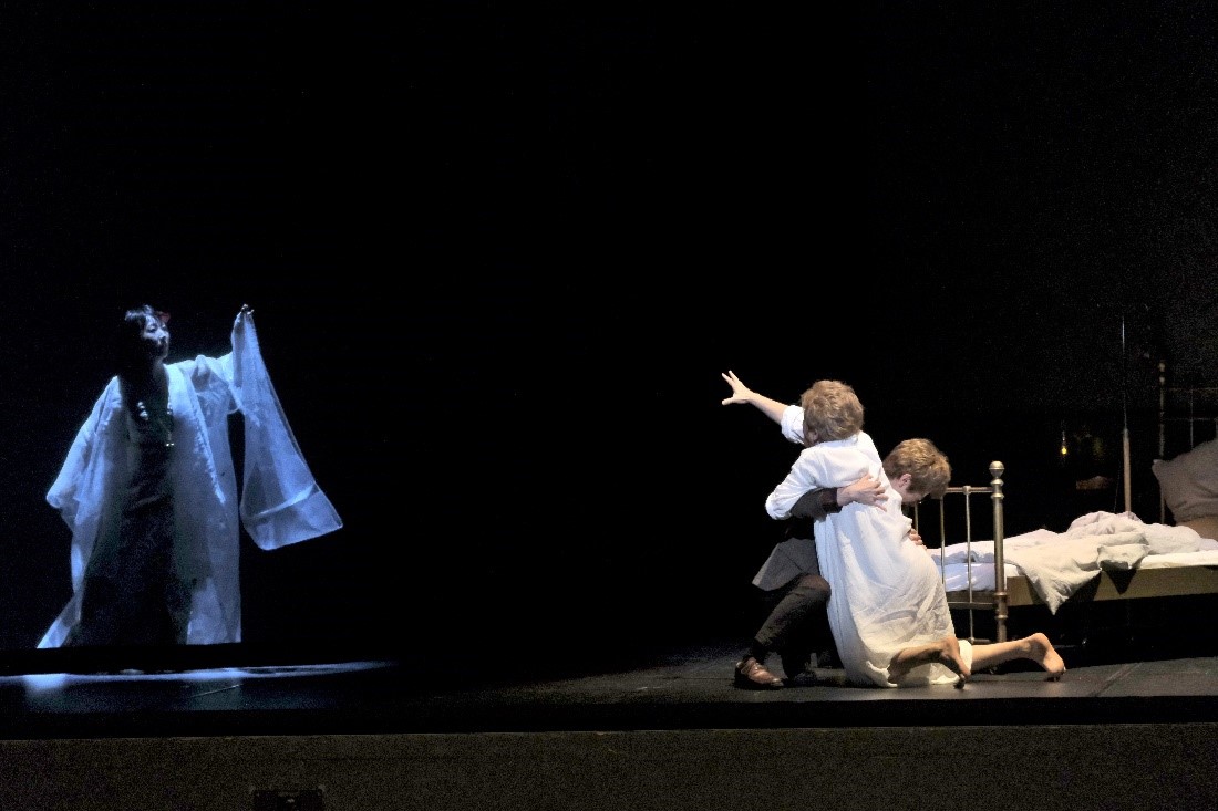 two people on stage clutch in desperation as what appears to be the spirit of a loved on attempts to communicate from further on the stage.