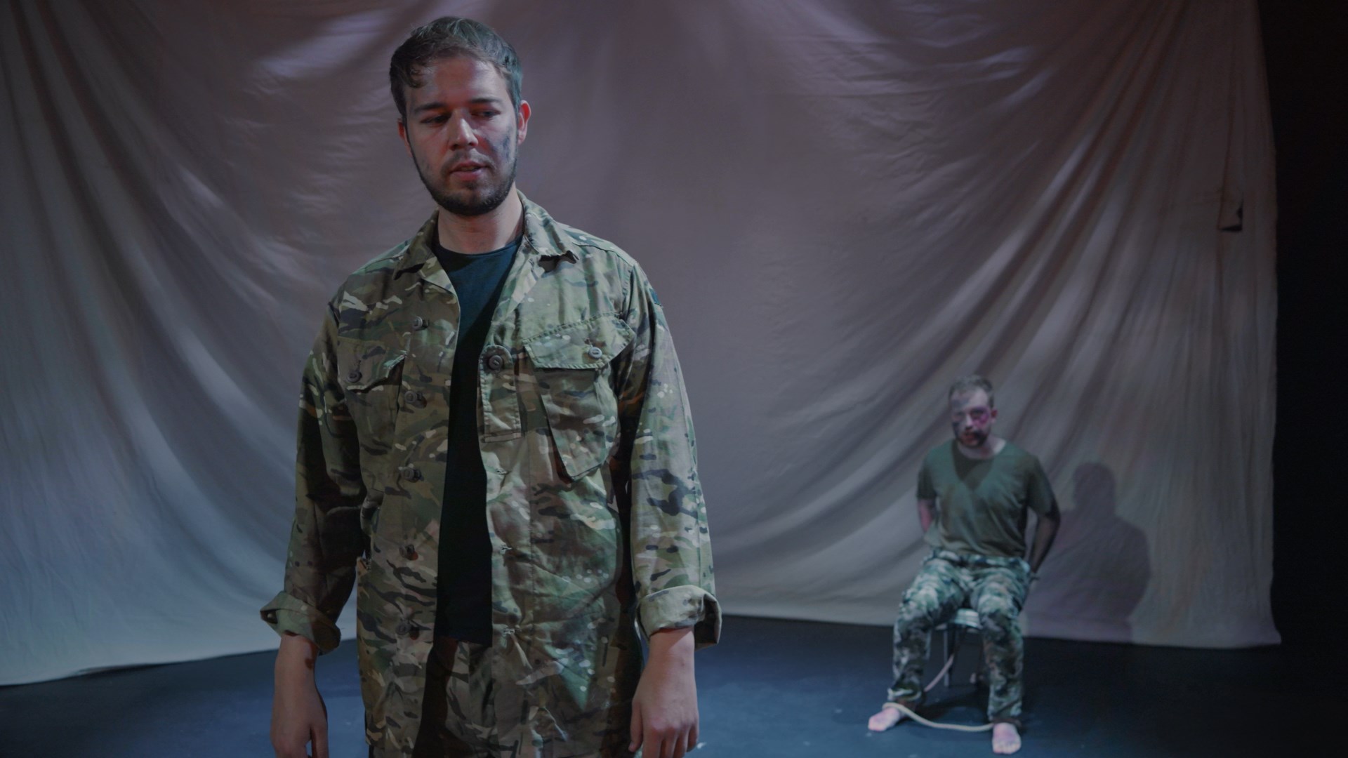 man in military fatigues with a prisoner behind him tied to a chair