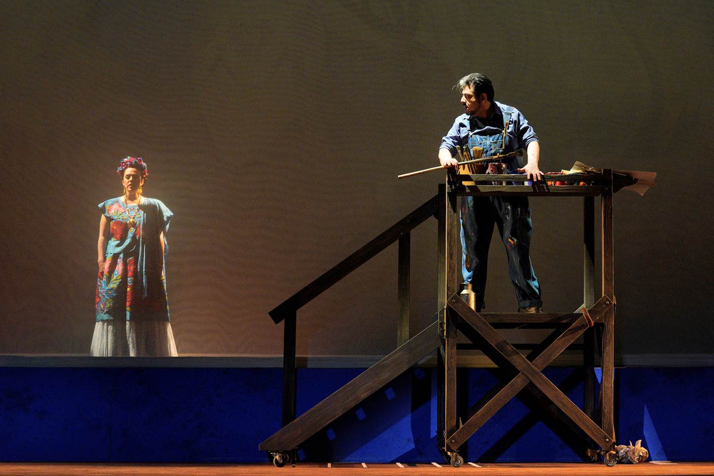 Scene from Frida of man in overalls with paint material standing on stairs looking at a woman in a colorful dress