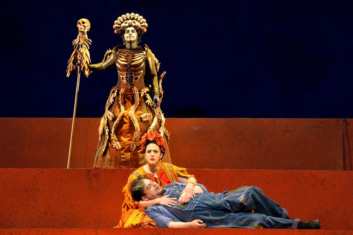 Scene from Frida of a woman holding a man laying on the floor while a person in a skeleton costume stands behind them.