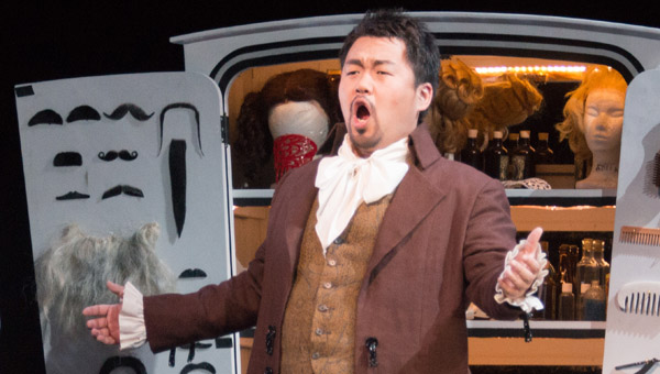 Barber of Seville singing in front of a wagon with wigs, mustaches and beards inside.
