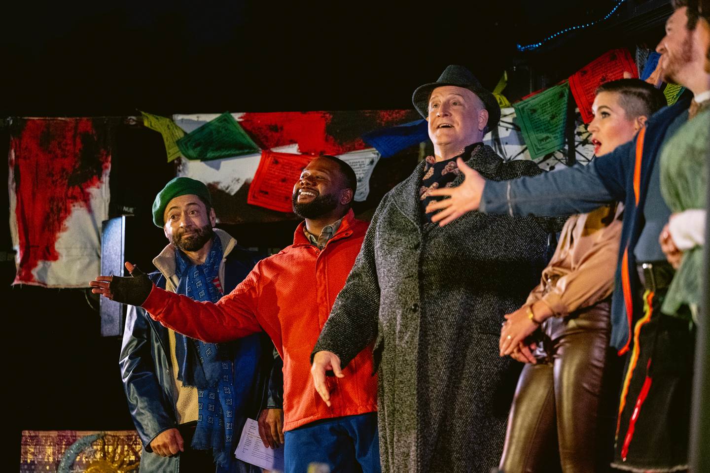 A vibrant group of five people is seen in a joyous pose, possibly in the midst of a theatrical performance. From left to right: a man in a green beanie and blue scarf extends his left arm out, palm up, with a facial expression of whimsical explanation. Next to him, a man in a bright red jacket smiles broadly, looking up and to his right with his left hand outstretched and fingers splayed. Beside him, an older gentleman in a gray hat and overcoat with a textured scarf gestures grandly with his right hand, as if emphasizing a point, his mouth open mid-speech. Next is a person with short hair and bold red lipstick, dressed in a brown coat and black pants with a multicolored stripe down the side; their right arm reaches out as if to grab someone&#39;s attention. Finally, a man on the far right wears a green jacket and scarf, with his hands clasped together in front of him. They stand before a background adorned with colorful prayer flags and various papers. The atmosphere is one of camaraderie and lively interaction.