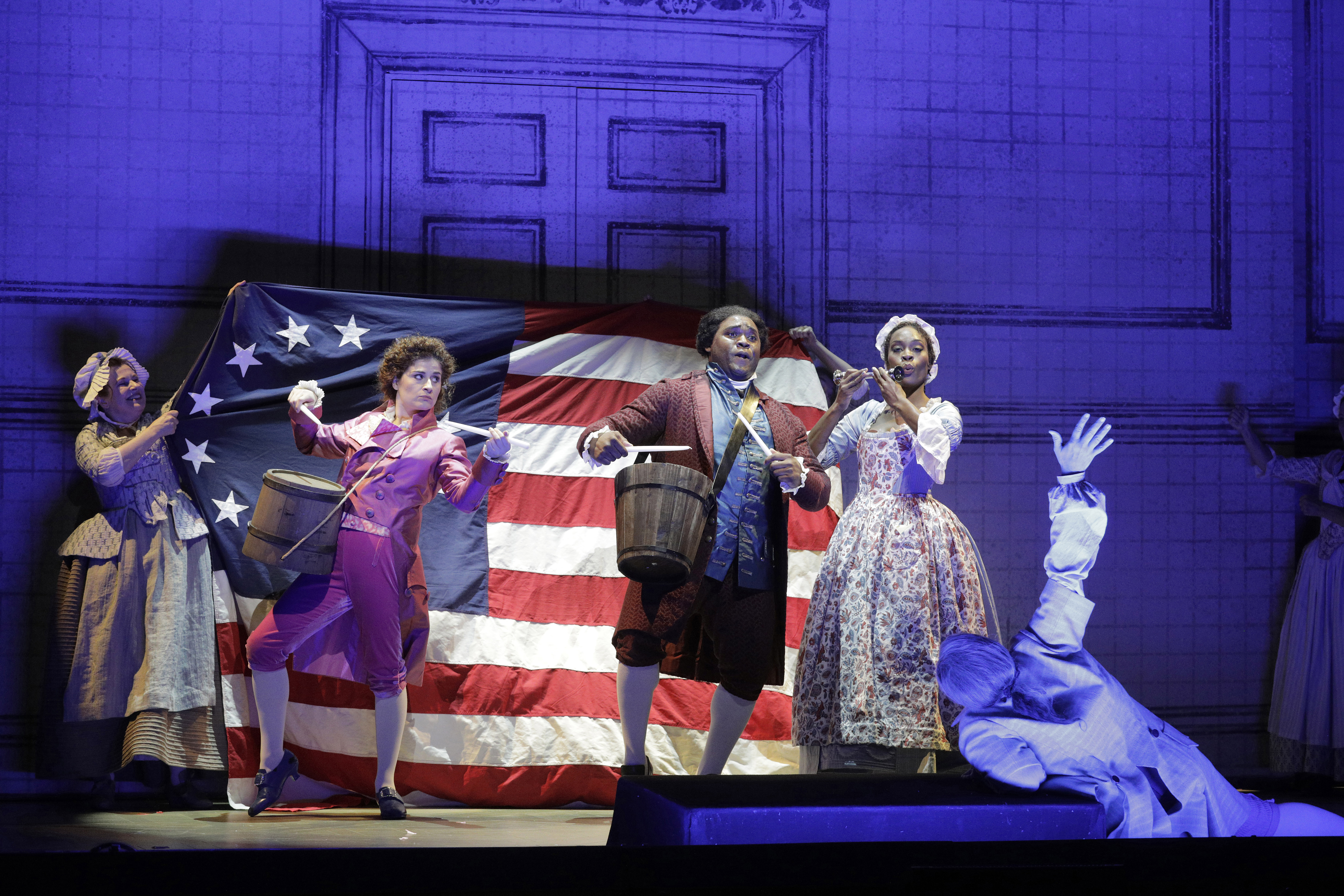 Performers in period costumes stand before an early American flag, with two playing drums and one person lying on the stage.