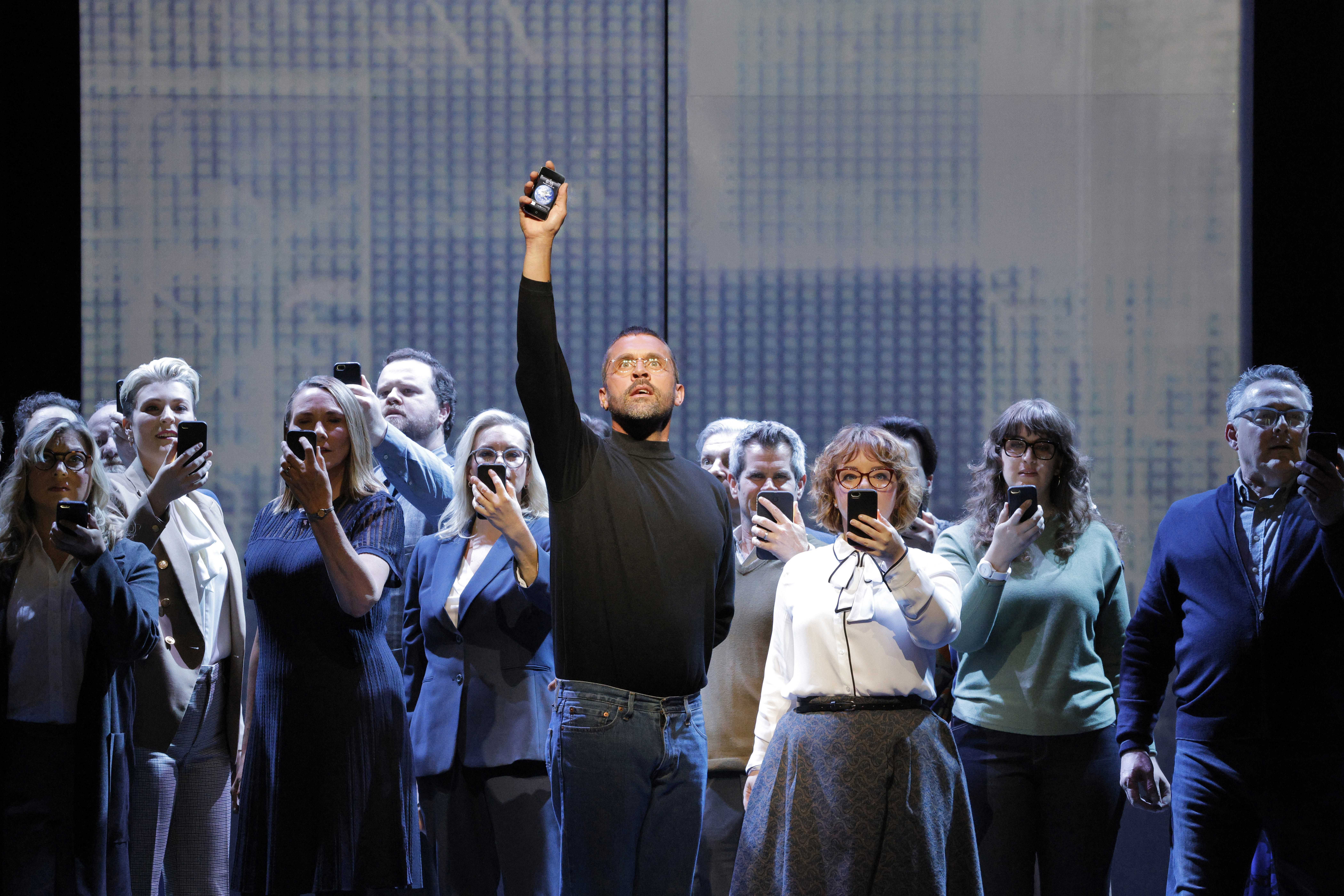 man holding iphone above his head while approximatel 15 other people stand behind him looking at their iphones