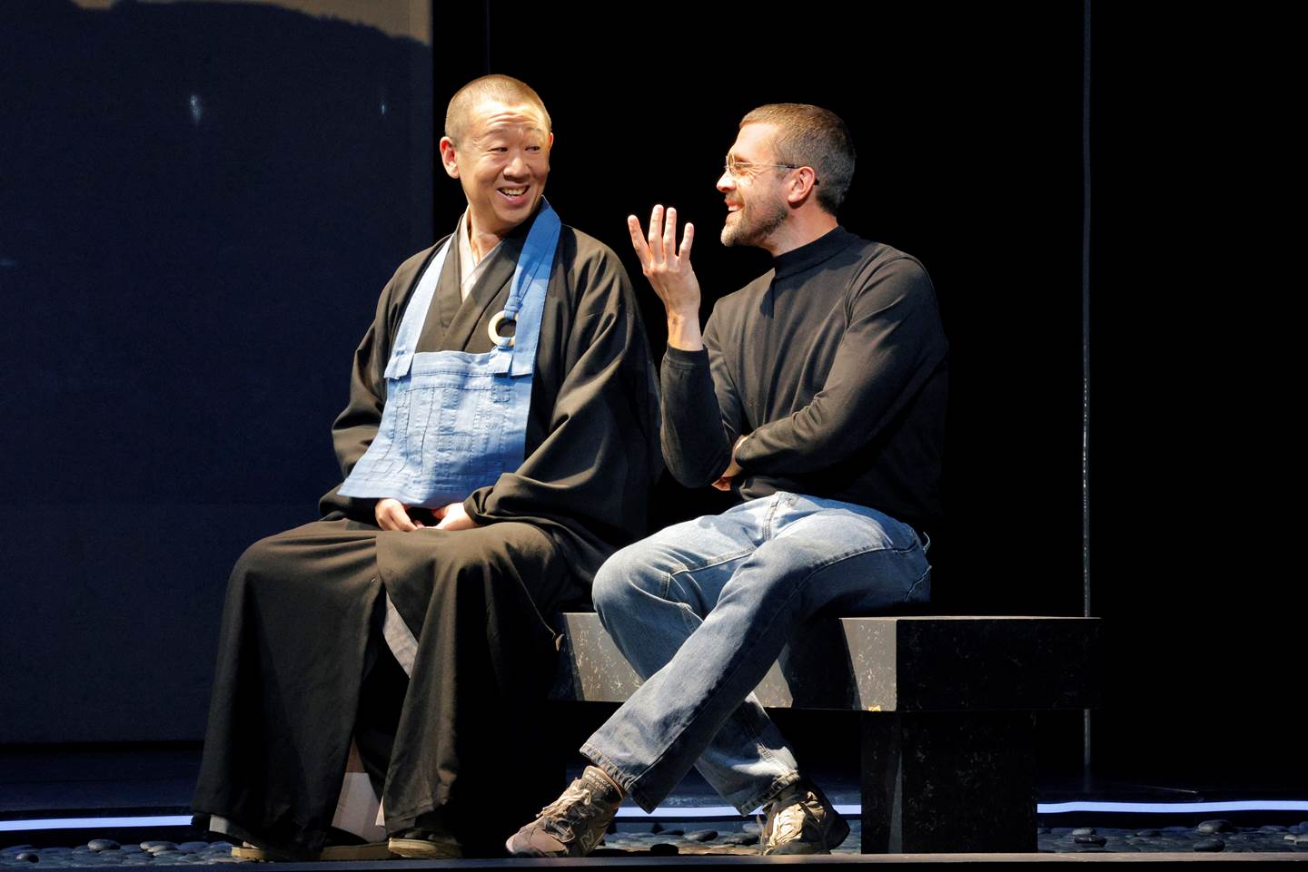 two men on stage talking and smiling while seated.