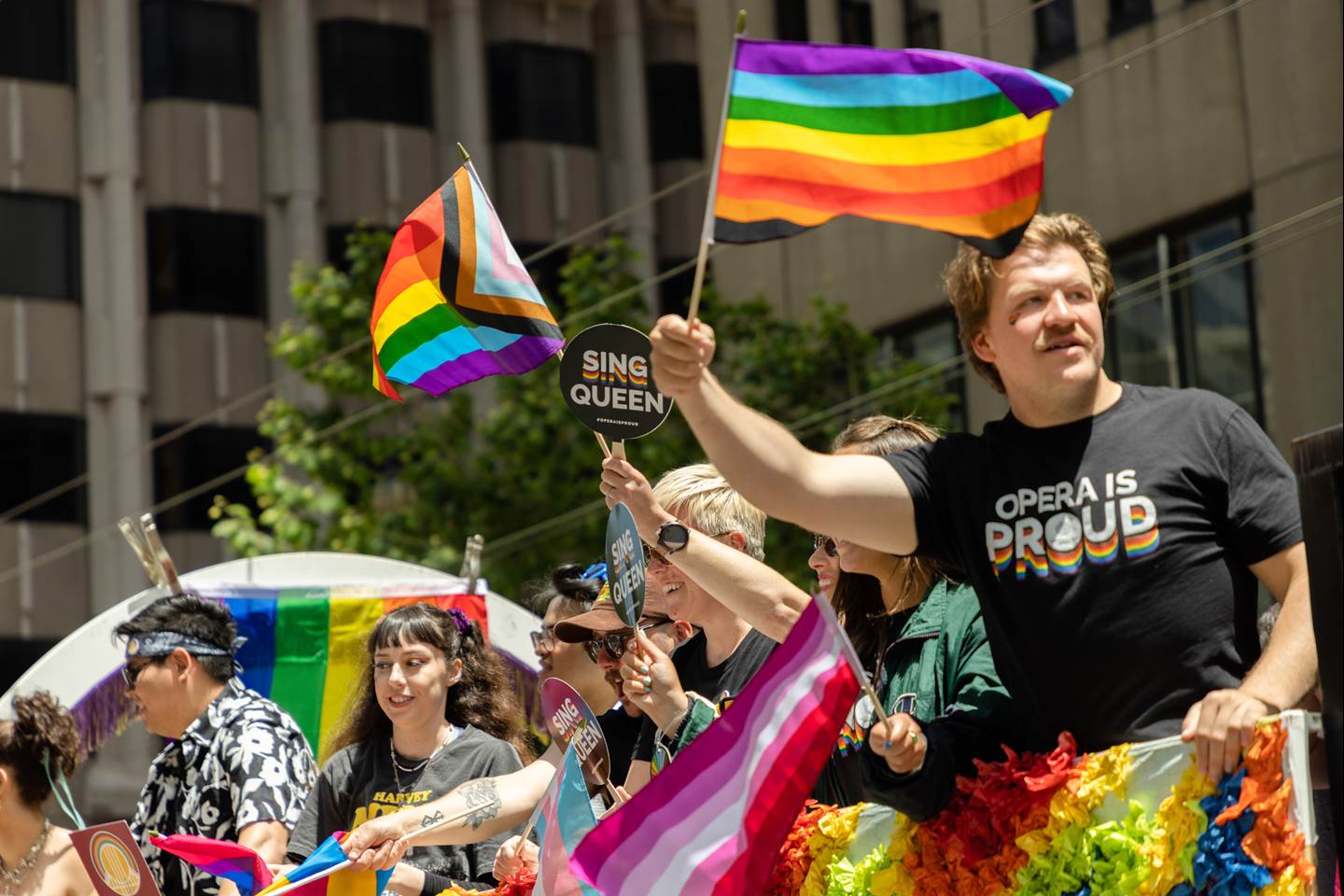 SFO cast members on the float waving colorful flags and signs that say &quot;Opera is Proud&quot; and &quot;Sing Queen&quot; in the 2023 Pride Parade.