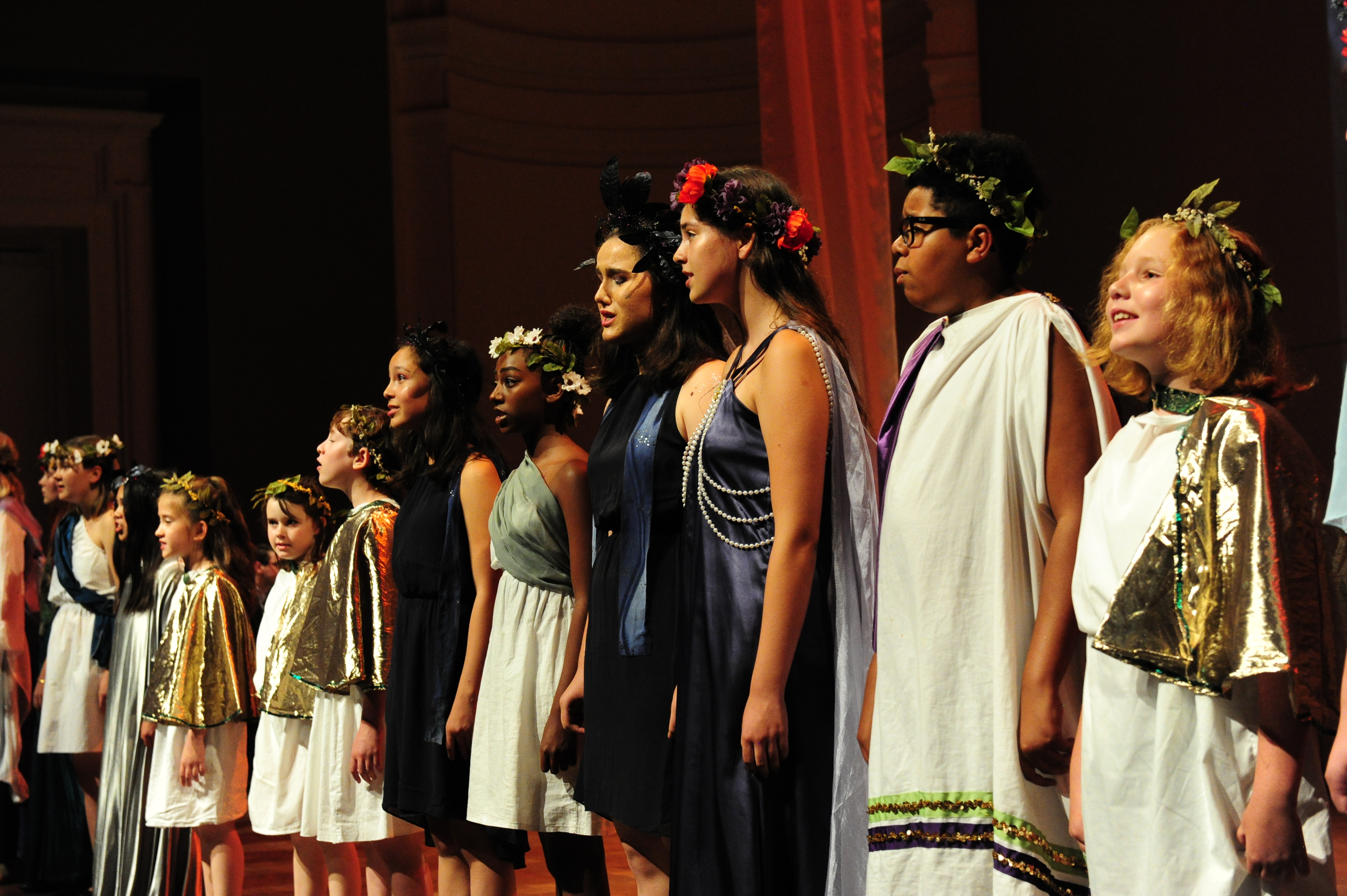 Group of children of all ages standing in a line on stage dressed in roman costumes